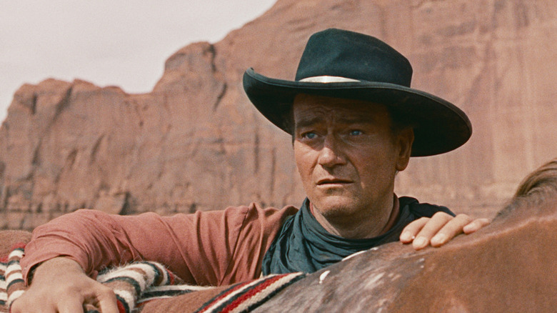 Ethan Edwards (John Wayne), wearing a black cowboy hat, looks intense and forlorn as he stands holding onto a horse's back against canyon cliffs in The Searchers