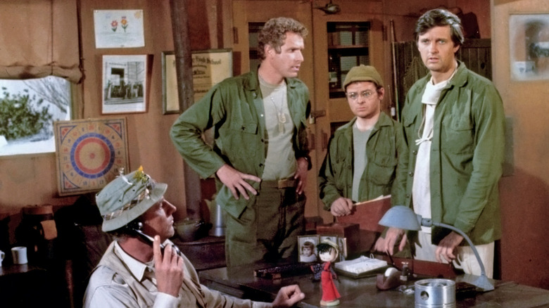 Trapper John (Wayne Rogers), Radar (Gary Burghoff), and Hawkeye (Alan Alda) stand next to a desk in an office while Henry (McLean Stevenson) is on the phone in M*A*S*H